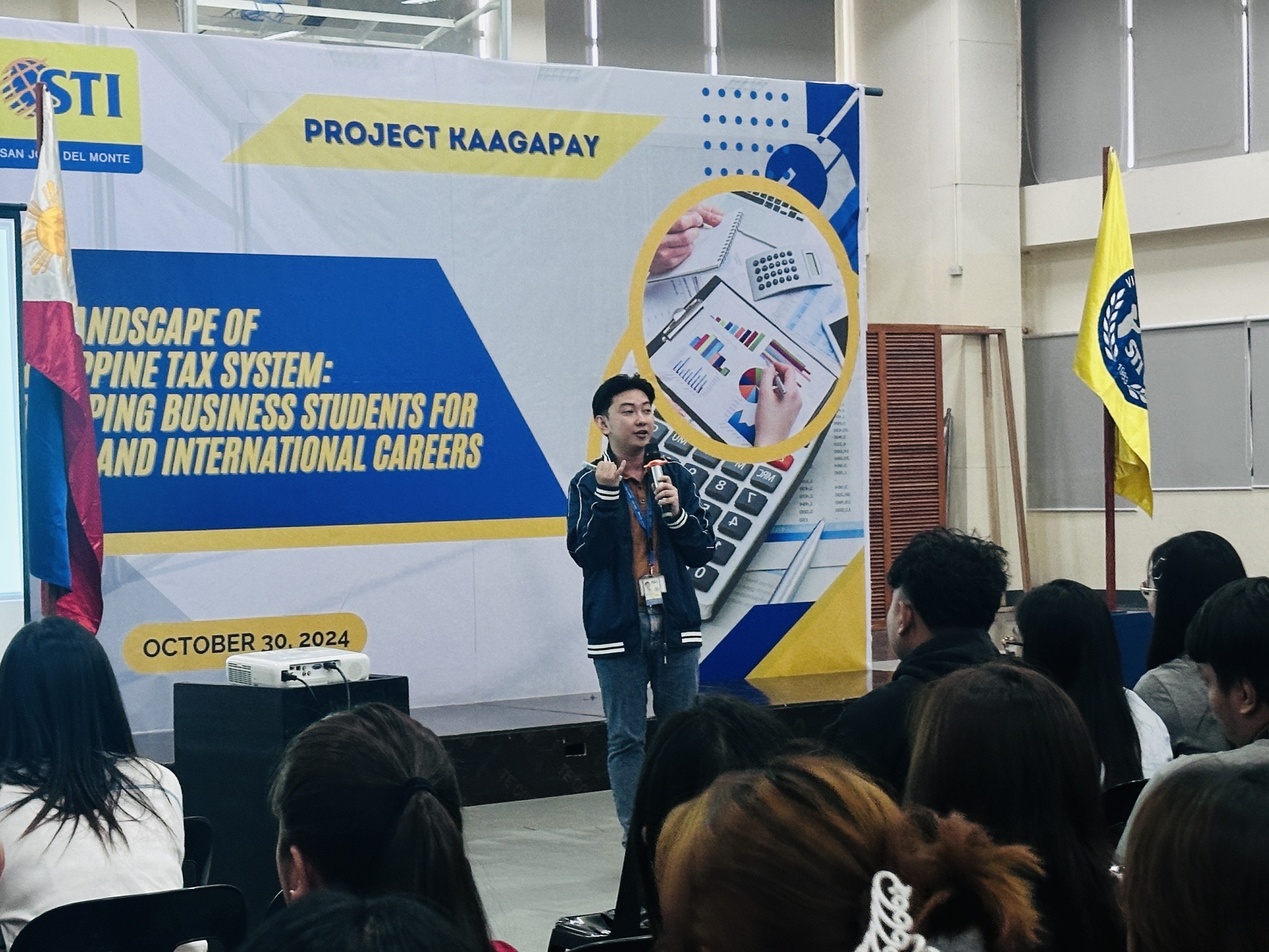 Jayson speaking with a microphone in front of the Project Kaagapay tax seminar banner at STI College